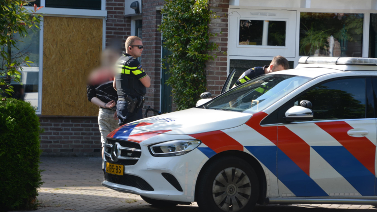 Twee politieagenten met een gearresteerde persoon naast een politieauto buiten een gebouw.