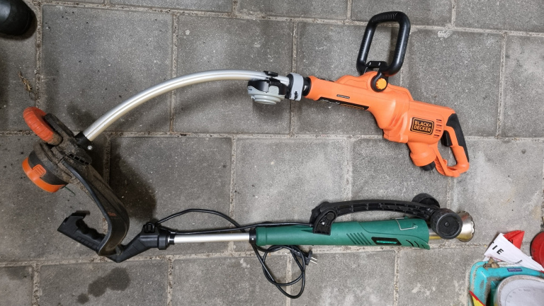 Twee elektrische grastrimmers op een stenen vloer, één oranje van Black & Decker en één groene met een elektriciteitskabel.