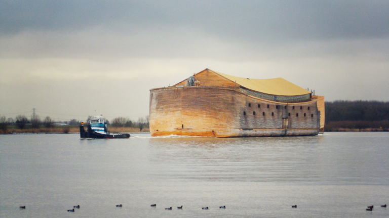 Houten ark op het water met een sleepboot ernaast onder een grijze lucht.