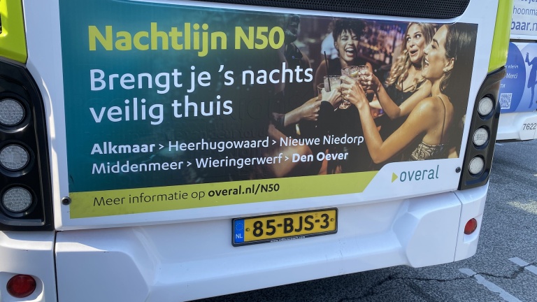 Poster op een bus met promotie voor Nachtlijn N50 die diverse steden verbindt; afbeelding van feestende mensen en een Nederlandse kentekenplaat (85-BJS-3) zichtbaar.
