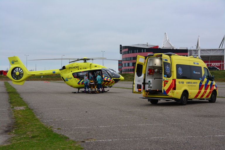 Een gele traumahelikopter en ambulance staan op een parkeerterrein; medisch personeel is bij de helikopter.