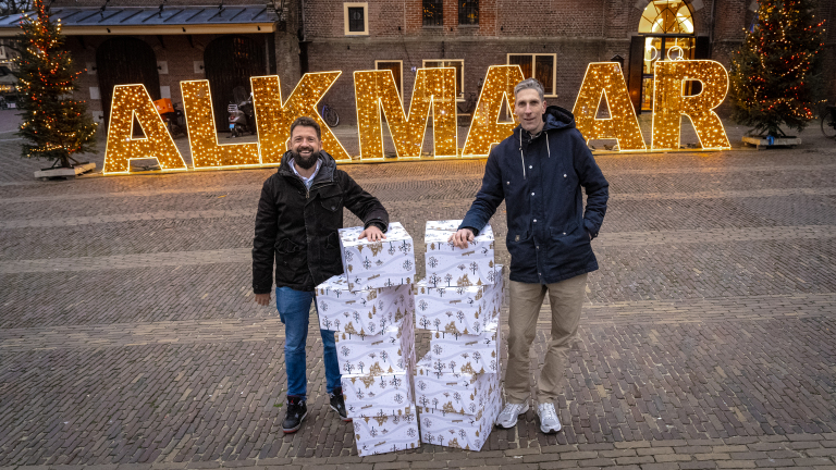 Twee mannen staan bij stapels ingepakte dozen met op de achtergrond verlichte letters "ALKMAAR".