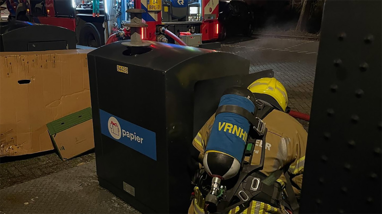 Brandweerman in beschermende uitrusting onderzoekt een papiercontainer naast een brandweerwagen.