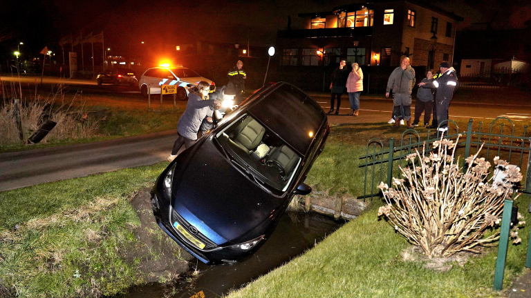 Auto in sloot met mensen eromheen en politie op de achtergrond bij avond.
