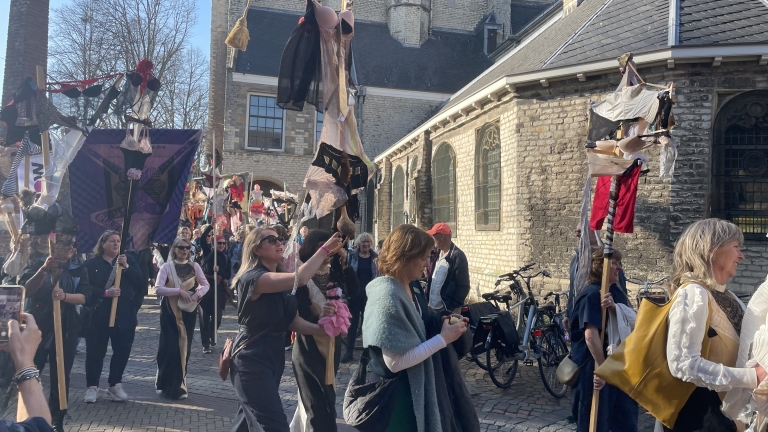 Een groep mensen loopt door een straat met historische gebouwen, terwijl ze stokken dragen versierd met kleurrijke kledingstukken.