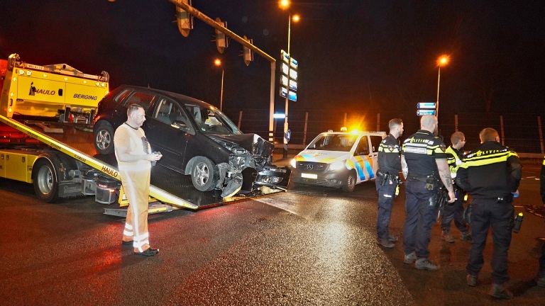 Auto met schade op een sleepwagen, omringd door politieagenten en een politiewagen, 's nachts bij een verlichte verkeerskruising.