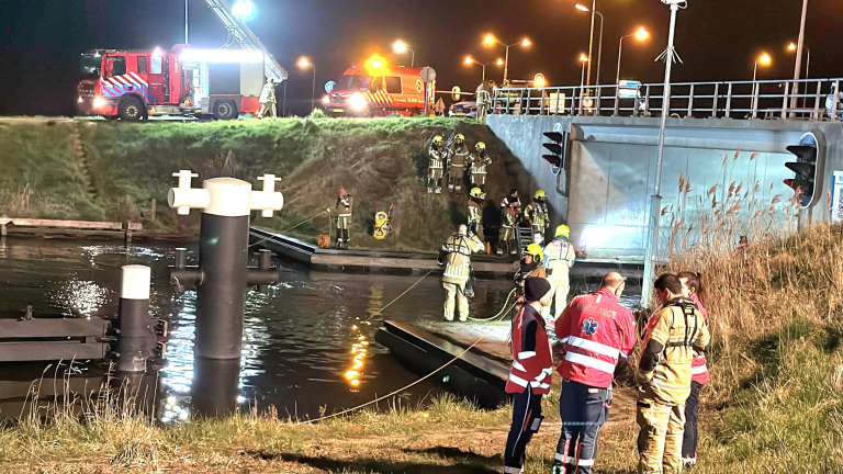 Brandweer en hulpdiensten werken 's nachts bij een brug aan een reddingsoperatie boven een kanaal, met brandweerlieden in uitrusting en voertuigen met zwaailichten.