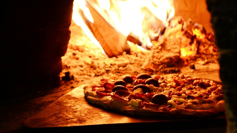 Pizza in een houtgestookte oven, met een vlam op de achtergrond.