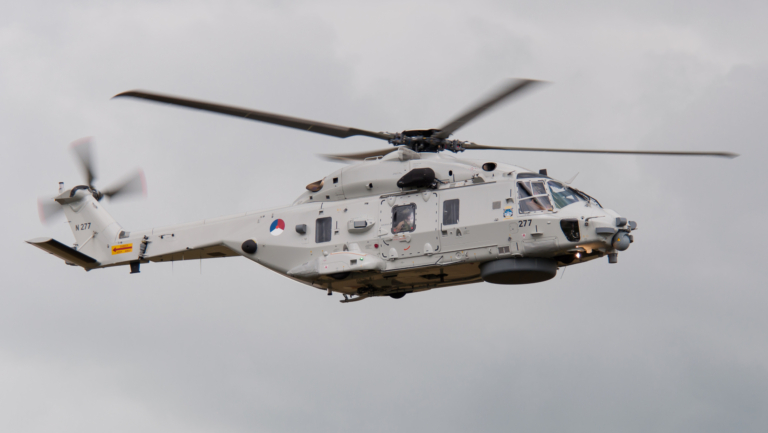 Grijze militaire helikopter in de lucht met draaiende rotors, boven een bewolkte hemel.