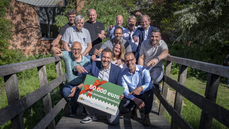Groep mensen poseert lachend op een brugje met een groot bord waarop "60.000 euro voor energiehub Beverkoog" staat, tegen een achtergrond van groen en een bakstenen gebouw.
