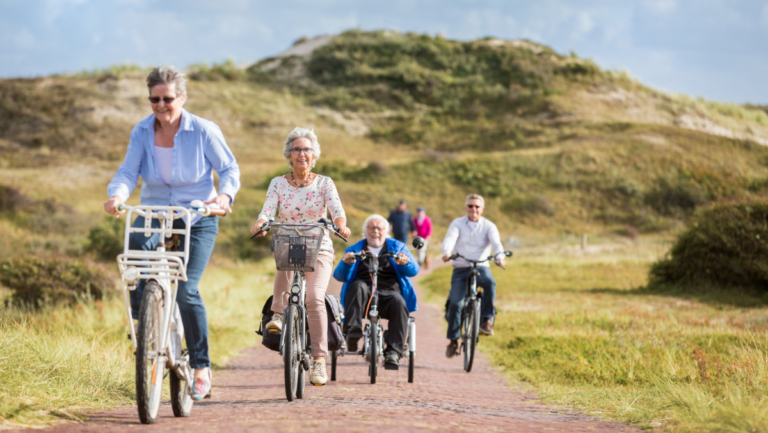 Vier oudere mensen fietsen over een pad door duinen op een zonnige dag.