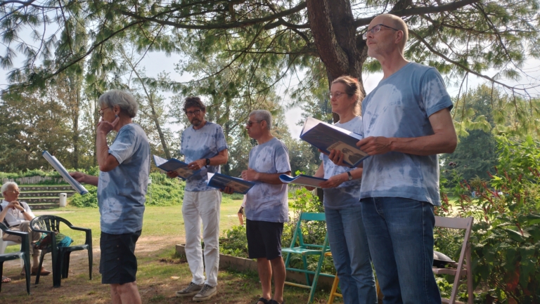 Groep mensen in blauwe shirts met muziekboekjes zingt buiten onder een boom.