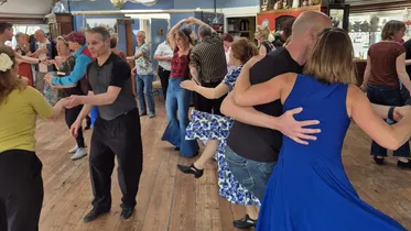 Groep mensen danst samen in een lichte zaal met houten vloer en grote ramen, sommige paren zijn in elegante houdingen tijdens een dansles of sociale dans.