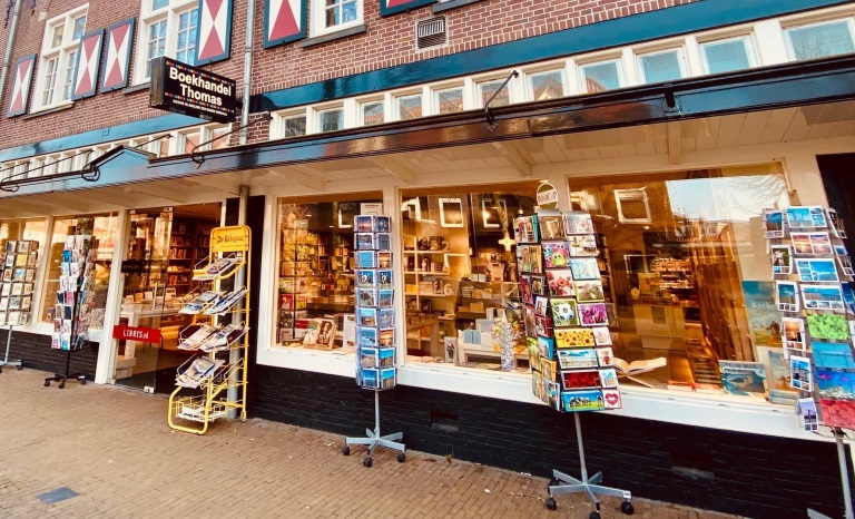 Buitenkant van een boekwinkel genaamd 'Boekhandel Thomas' met verschillende stellingen vol ansichtkaarten voor de etalage.