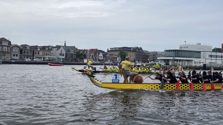 Drakenbotenrace met twee gele boten met drakenkoppen voorop, teams van mensen in gele en zwarte shirts peddelen synchroon, op een rivier met stadsgebouwen op de achtergrond.
