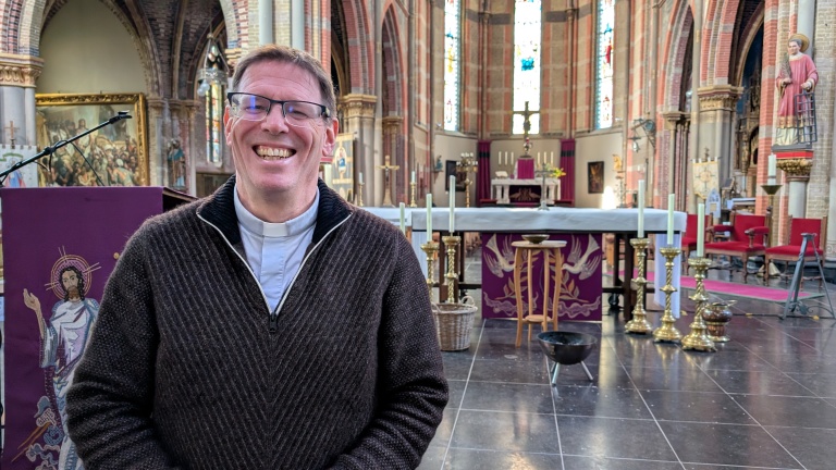 Een lachende man met een priesterboord in een kerk met altaar en versierde kaarsen op de achtergrond.