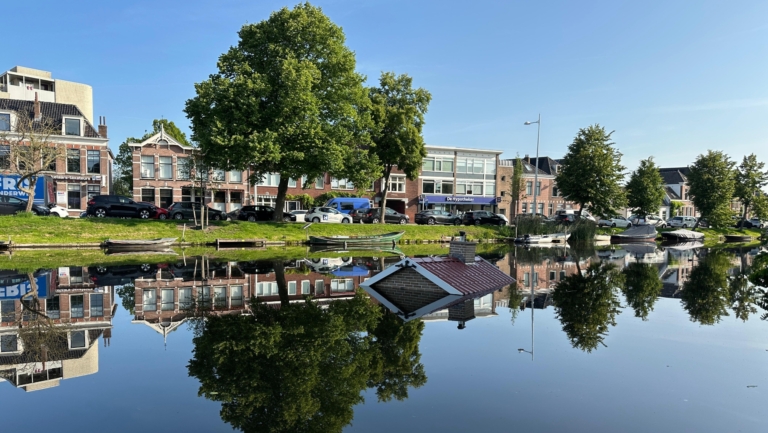 Gracht met huizen en bomen erlangs, met een dak van een huis dat onder water lijkt te staan, alles weerspiegeld in het kalme wateroppervlak.