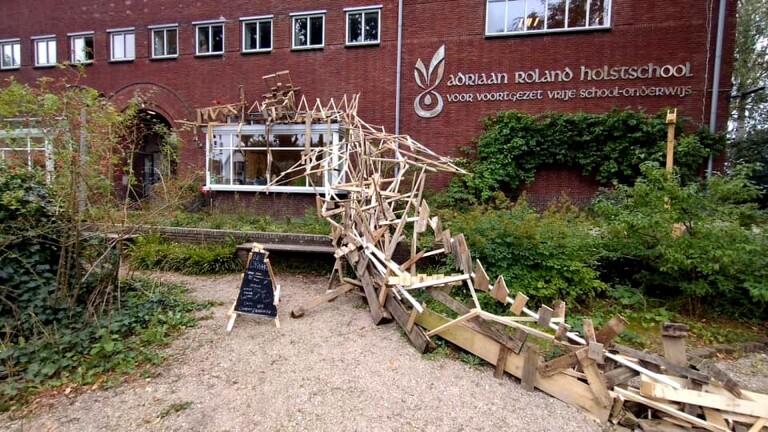 Buiten kunstinstallatie van houten planken in organische vormen voor de Adriaan Roland Holstschool, met groene begroeiing en een schoolbord ernaast.