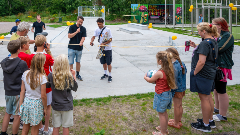 Een ceremonieel openingsmoment van een skatepark, met een groep volwassenen en kinderen die toekijken terwijl twee mannen een lint doorknippen. Een van de mannen heeft een skateboard en een microfoon in de hand. De omgeving is versierd met gele ballonnen en er zijn betonnen skatevoorzieningen zichtbaar. Op de achtergrond zijn bomen en een kleurrijke muurschildering te zien.