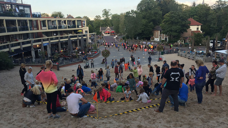 Groep mensen, waaronder veel kinderen, verzameld op een zandhelling voor een evenement, met een gebouw en terras op de achtergrond en een straat omringd door bomen verder weg.