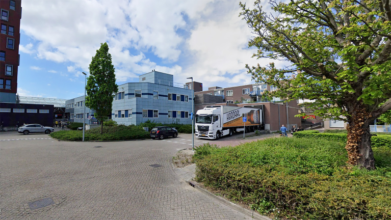 Straatbeeld met een vrachtwagen geparkeerd naast een modern gebouw met blauwe tegels, omgeven door groen en bomen, onder een bewolkte hemel.