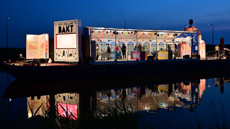 Een verlichte opstelling van "Heel Holland Bakt" aan het water in de avond; het heeft een open keuken met mensen binnen, versierd met lichtjes en kleurrijke decoraties.
