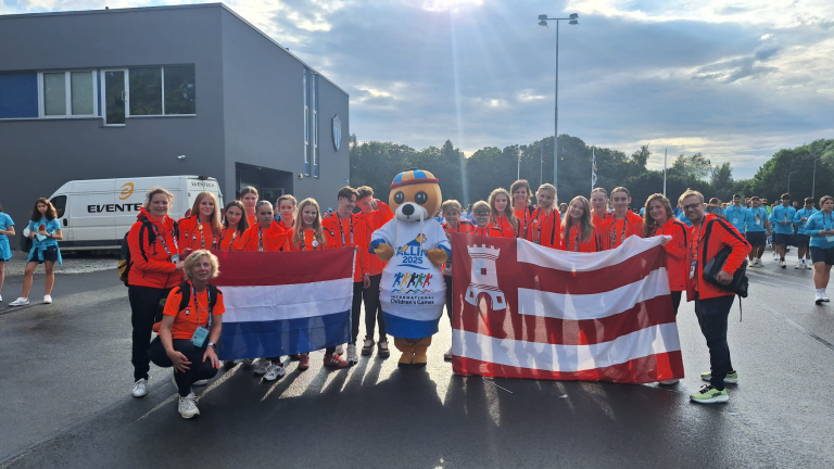 Een groep mensen in oranje sportkleding poseert met twee vlaggen naast een mascotte in een T-shirt met de tekst 'KLIN 2025 International Children's Games'.