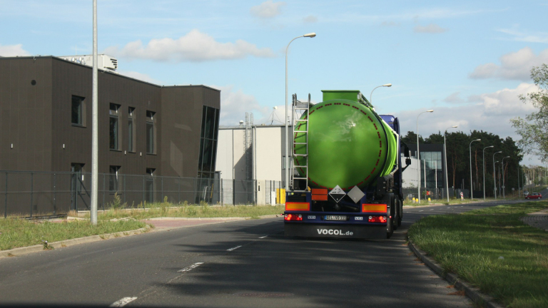 Vrachtwagen met een grote, groene tank rijdt over een weg naast een modern industrieel gebouw met een donkere gevel. Lantaarnpalen zijn te zien langs de weg, met bomen en gras ernaast.