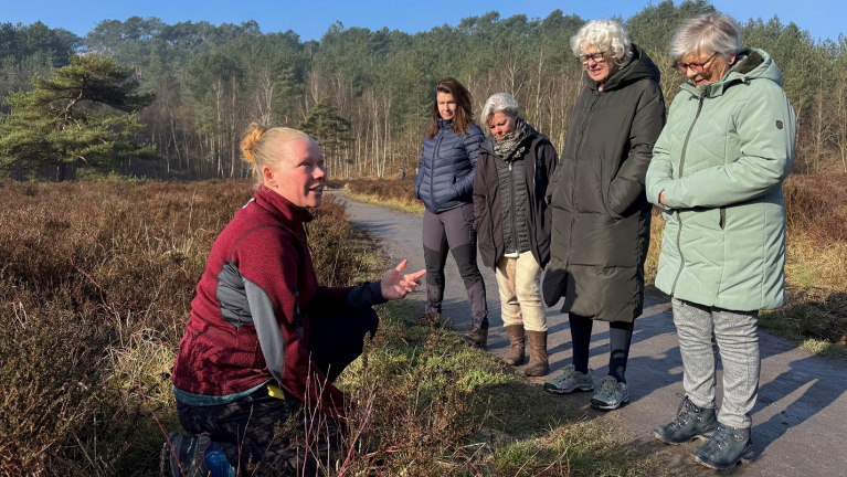 Een vrouw in een rood en grijs jack hurkt voor een groep van vier vrouwen tijdens een wandeling in een bosrijk gebied onder een zonnige hemel.