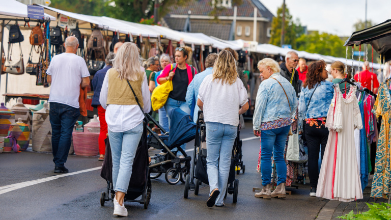 Mensen lopen over een drukke marktplaats met kramen vol handtassen en kleding, met enkele personen die kinderwagens duwen.
