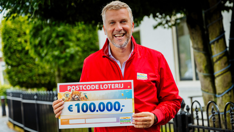 Man in a red jacket smiling while holding a €100,000 Postcode Loterij sign.