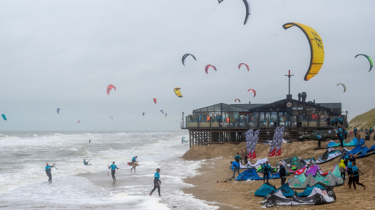 Kitesurfers op een strand bij een houten strandpaviljoen, met meerdere kites in de lucht en mensen die voorbereidingen treffen op het zand.