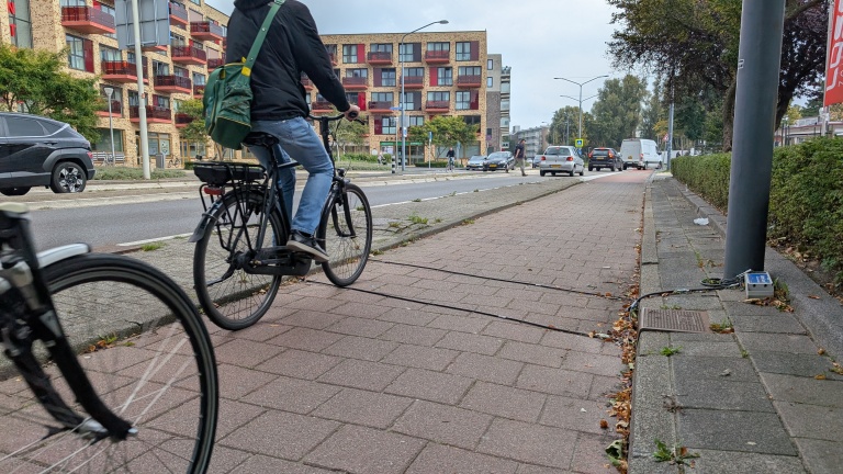 Een fietspad met een fietser op de voorgrond, in de buurt van een drukke straat in een stedelijke omgeving. Er zijn kabels en een meetapparaat aan de rechterkant van het pad.