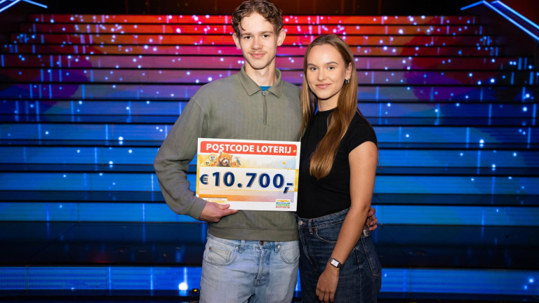 Een jong stel poseert met een cheque van de Postcode Loterij ter waarde van €10.700 in een kleurrijke en verlichte omgeving.