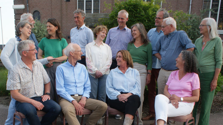 Een groep van dertien mensen, zowel zittend als staand, lachend en pratend op een buitenlocatie voor een gebouw. Ze dragen casual kleding in verschillende kleuren.