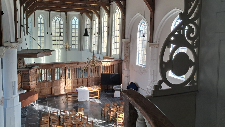 Interieur van een historische kerk met hoge glas-in-loodramen, een houten preekstoel, en rijtjes houten stoelen gericht naar een centrale tafel met kandelaars en een wit kleed.