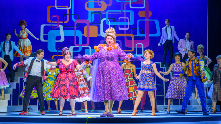 Theaterproductie met een groep acteurs in kleurrijke jaren '60 kleding op een podium met een geometrisch patroon op de achtergrond. De centrale acteur draagt een glinsterende paarse jurk met bloemen en handschoenen, omringd door dansende en lachende medespelers.