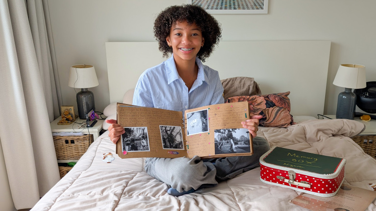 Jonge persoon in een blauwe blouse zit gekruist op een bed en houdt een open scrapbook vast met foto's en geschreven teksten erin. Naast hen ligt een rode koffer met witte stippen en de tekst "Memory Box" erop. Op de achtergrond is een slaapkamer te zien met lampen en een nachtkastje.