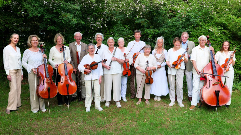 Groep musici in witte en beige kleding poseren op een grasveld met hun instrumenten, waaronder cello's, een contrabas, een hobo en violen, tegen een achtergrond van groene struiken.
