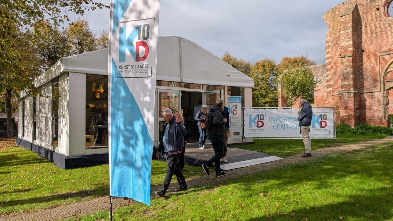 Mensen lopen naar een informatiecentrum voor Kunst 10 Daagse Bergen op een grasveld met bomen en een bakstenen gebouw op de achtergrond.