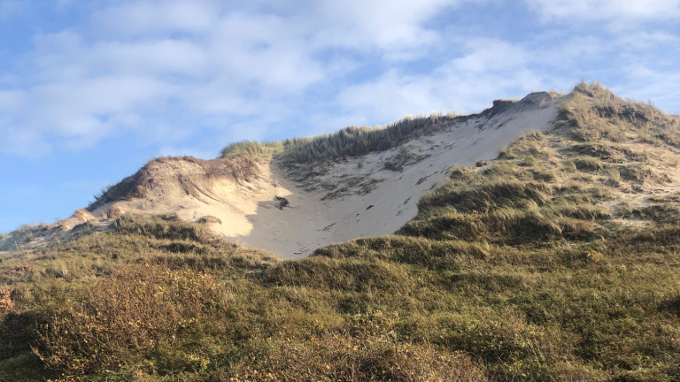Zandduin met gras bedekte helling onder een blauwe lucht met enkele wolken.