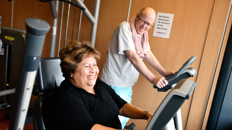 Twee mensen sporten in een fitnessruimte; een vrouw op een stationaire fiets en een man op een loopband lachen samen.