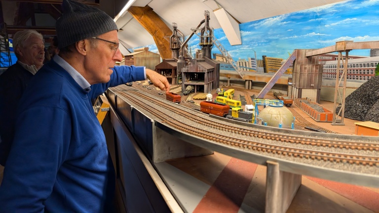 Een man in een blauwe trui en grijze muts bedient een modeltreinbaan met industriële gebouwen op de achtergrond, terwijl een oudere man toekijkt.