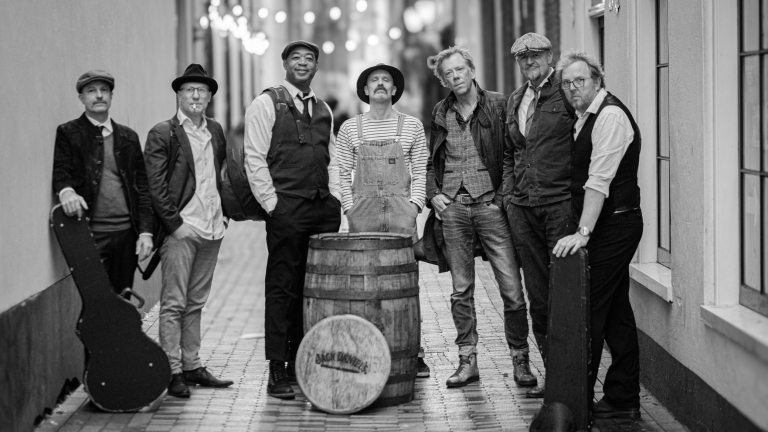 Zeven mannen in vintage kleding staan in een smalle straat rond een houten vat, waarvan de voorzijde een Jack Daniel's logo toont.