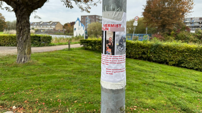 Een vermist poster is geplakt op een lantaarnpaal in een groene buitenomgeving. Op de poster staan afbeeldingen van een persoon en een fiets, met daarin tekst over een vermissing, contactinformatie en een oproep voor informatie.