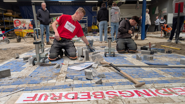 Twee mannen knielen op een bouwplaats binnen een werkplaats en leggen bestrating met stenen en zand. Er liggen gereedschappen zoals een schop en een steenknipper op de grond. Op de achtergrond staan enkele toeschouwers en andere arbeiders.