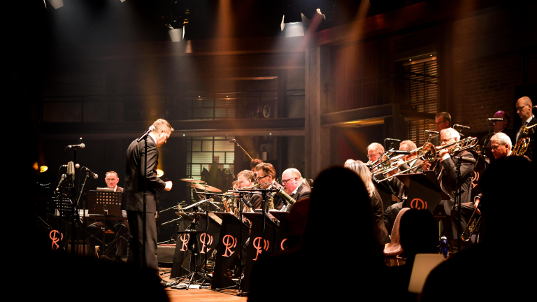 Een orkest speelt op een verlicht podium, met een dirigent die voor de muzikanten staat. De blazerssectie zit prominent aan de rechterkant.