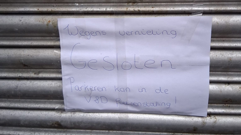 Een handgeschreven briefje op een metalen garagedeur met de tekst: 'Wegens vernieling gesloten. Parkeren kan in de V&D fietsenstalling!'