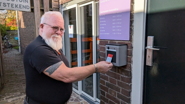 Een oudere man met een witte baard en bril gebruikt een pas bij een toegangscontrole bij een fietsparkeerplaats. Achter hem zijn borden met openingstijden zichtbaar.