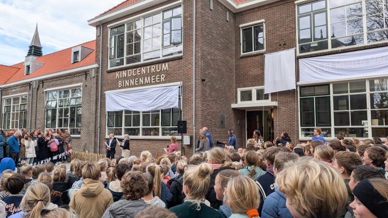 Grote groep mensen, voornamelijk kinderen, verzameld voor het Kindcentrum Binnenmeer, een bakstenen gebouw met grote ramen en een rood dak, tijdens een evenement.